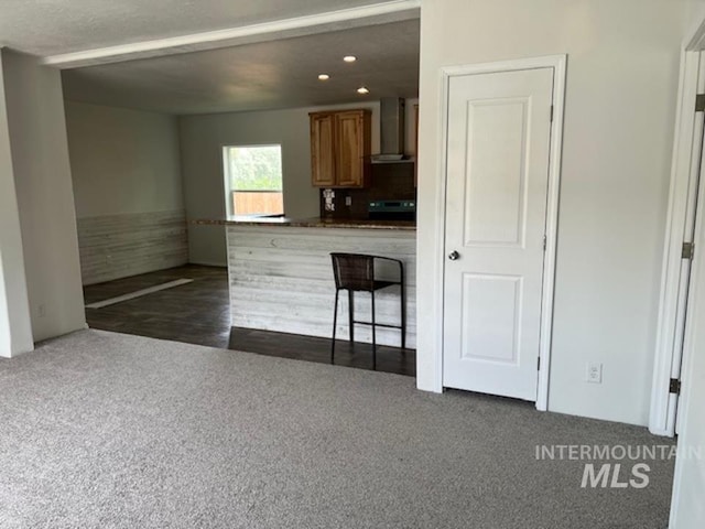 kitchen featuring wall chimney range hood, brown cabinets, dark colored carpet, dark countertops, and recessed lighting