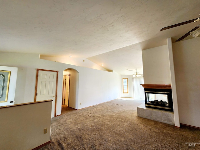 unfurnished living room featuring ceiling fan with notable chandelier, a tiled fireplace, vaulted ceiling, and carpet flooring