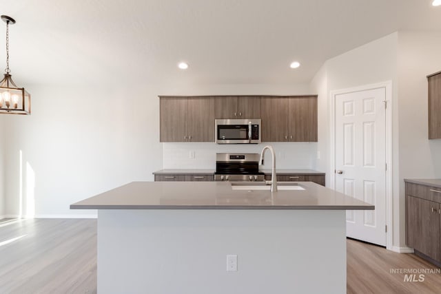 kitchen with a kitchen island with sink, a sink, light wood-style floors, appliances with stainless steel finishes, and a chandelier