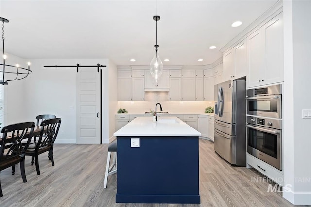 kitchen with white cabinetry, hanging light fixtures, stainless steel appliances, a barn door, and an island with sink