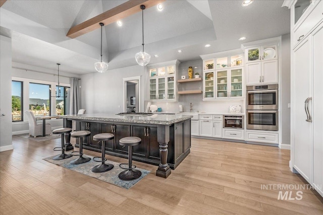 kitchen featuring a breakfast bar, white cabinets, an island with sink, double oven, and decorative light fixtures