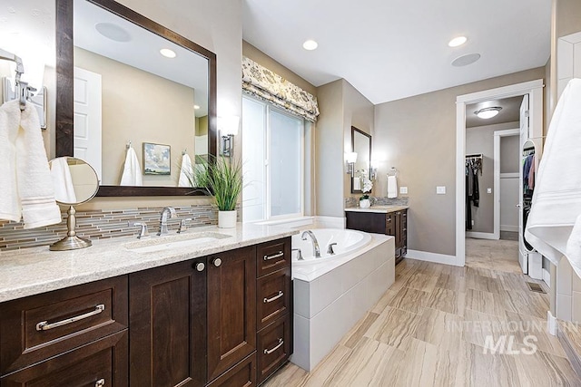 full bathroom with a walk in closet, a garden tub, two vanities, a sink, and backsplash