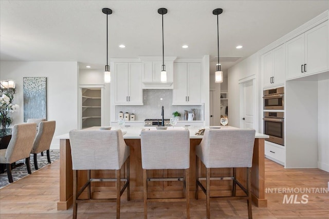 kitchen with white cabinets, light hardwood / wood-style floors, pendant lighting, decorative backsplash, and a kitchen island with sink