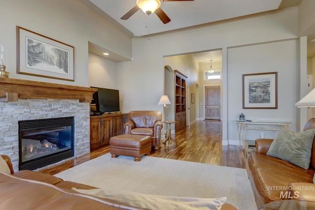 living area with ceiling fan, a stone fireplace, wood finished floors, and baseboards