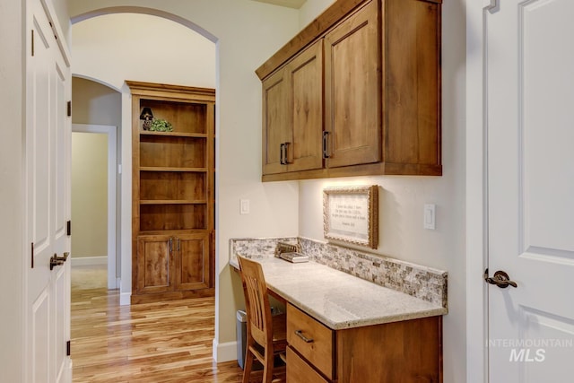 kitchen featuring arched walkways, brown cabinetry, light wood-style floors, light stone countertops, and baseboards