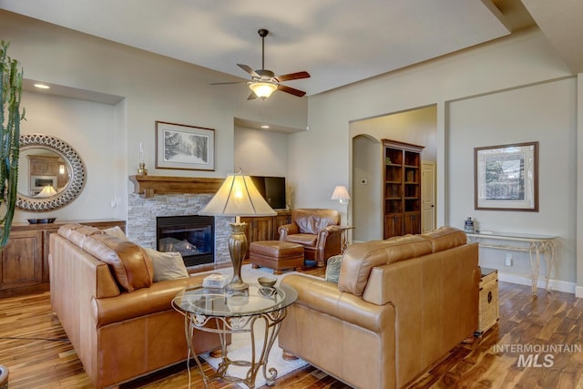 living area featuring ceiling fan, baseboards, wood finished floors, and a stone fireplace