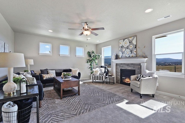 carpeted living room with a mountain view, ceiling fan, a stone fireplace, and a textured ceiling