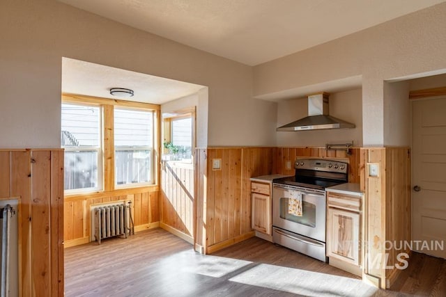 kitchen with wooden walls, stainless steel electric range oven, radiator heating unit, light countertops, and wall chimney range hood