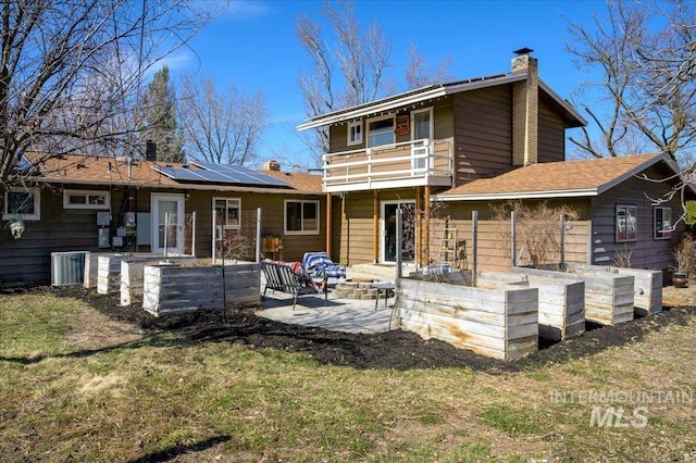 rear view of house with roof mounted solar panels, a patio, a yard, a balcony, and a chimney