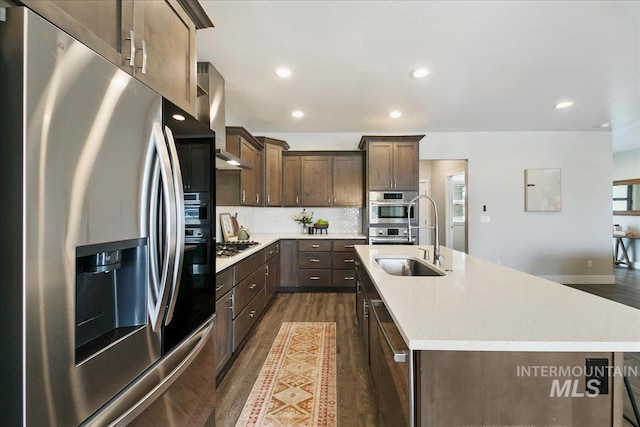 kitchen with dark wood-type flooring, a center island with sink, a sink, appliances with stainless steel finishes, and wall chimney exhaust hood