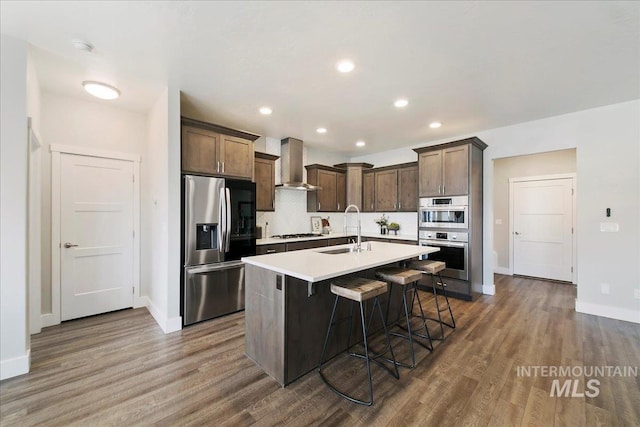 kitchen with wall chimney range hood, light countertops, appliances with stainless steel finishes, wood finished floors, and a sink