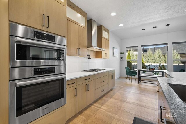 kitchen with wall chimney range hood, hanging light fixtures, light hardwood / wood-style flooring, a textured ceiling, and stainless steel appliances