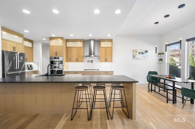 kitchen with dark stone counters, stainless steel appliances, wall chimney range hood, light hardwood / wood-style floors, and hanging light fixtures