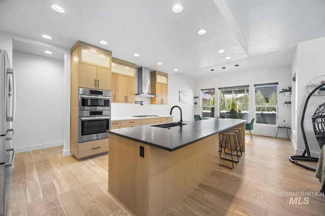 kitchen with sink, wall chimney range hood, light hardwood / wood-style floors, a center island with sink, and appliances with stainless steel finishes