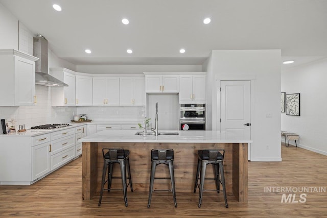 kitchen featuring a kitchen island with sink, white cabinets, a kitchen breakfast bar, wall chimney range hood, and sink