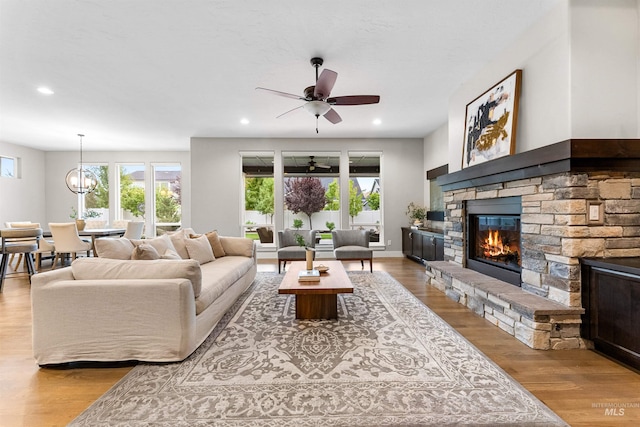 living area with light wood-style flooring, recessed lighting, a stone fireplace, and ceiling fan with notable chandelier