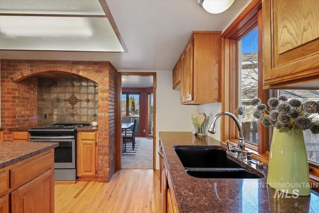 kitchen with stainless steel range, sink, a healthy amount of sunlight, light hardwood / wood-style flooring, and dark stone countertops