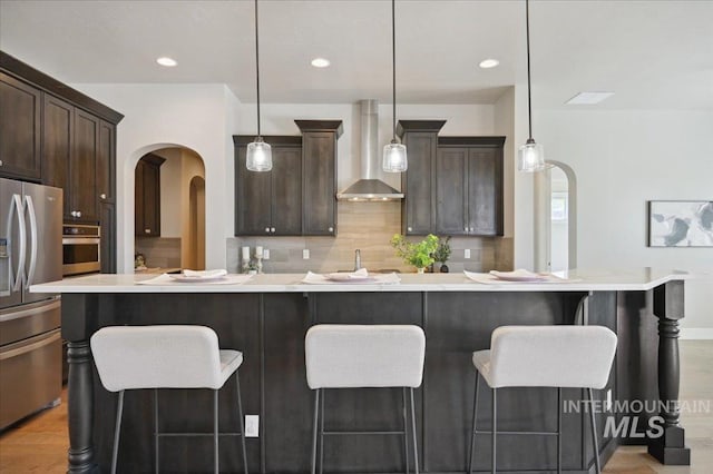 kitchen featuring stainless steel appliances, arched walkways, dark brown cabinets, decorative backsplash, and wall chimney range hood