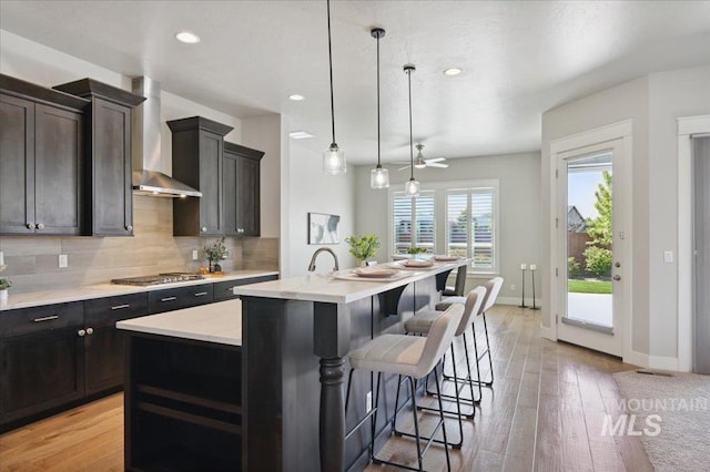 kitchen with wall chimney range hood, a kitchen breakfast bar, stainless steel gas stovetop, decorative backsplash, and an island with sink
