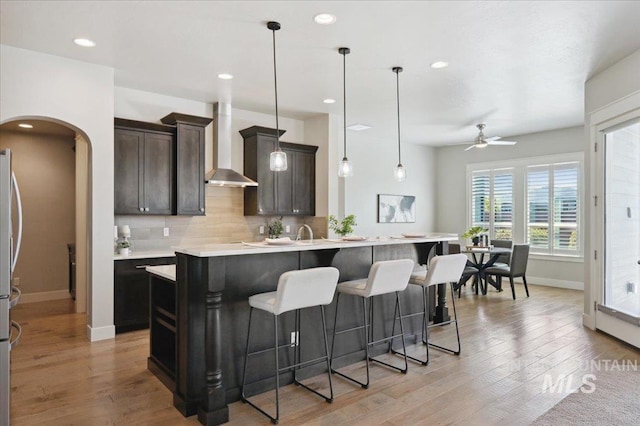 kitchen featuring wall chimney range hood, arched walkways, baseboards, a breakfast bar area, and ceiling fan