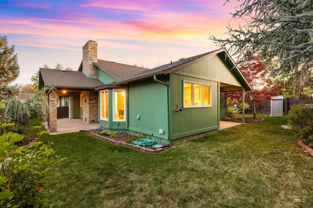 back of house at dusk featuring a chimney, fence, a shingled roof, a yard, and a patio