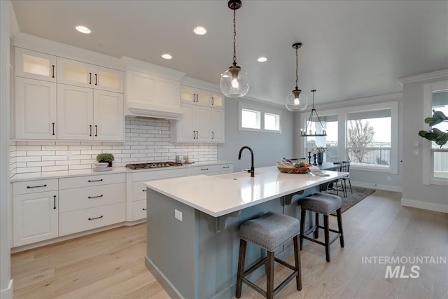 kitchen featuring a kitchen island with sink, light wood-type flooring, tasteful backsplash, decorative light fixtures, and white cabinetry