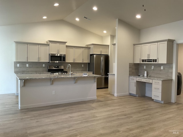 kitchen with light wood finished floors, a kitchen bar, light stone counters, stainless steel appliances, and a kitchen island with sink