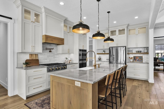 kitchen featuring stainless steel appliances, light wood-style flooring, decorative backsplash, a center island with sink, and decorative light fixtures