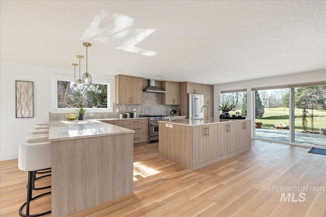 kitchen featuring light brown cabinets, light wood finished floors, appliances with stainless steel finishes, wall chimney range hood, and tasteful backsplash