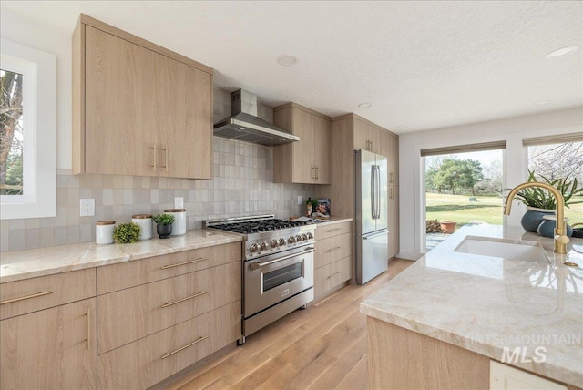 kitchen with a sink, appliances with stainless steel finishes, light brown cabinets, and wall chimney range hood