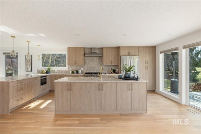 kitchen with light wood-type flooring, light brown cabinets, a sink, freestanding refrigerator, and wall chimney range hood