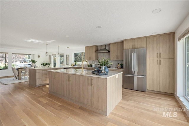 kitchen featuring a center island with sink, light brown cabinetry, freestanding refrigerator, wall chimney exhaust hood, and light wood finished floors