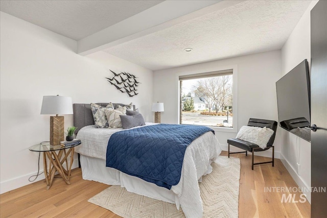 bedroom featuring light wood-type flooring, baseboards, a textured ceiling, and beamed ceiling