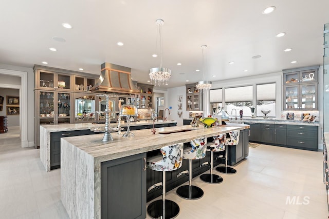 kitchen with a large island with sink, wall chimney range hood, gray cabinets, and light stone countertops