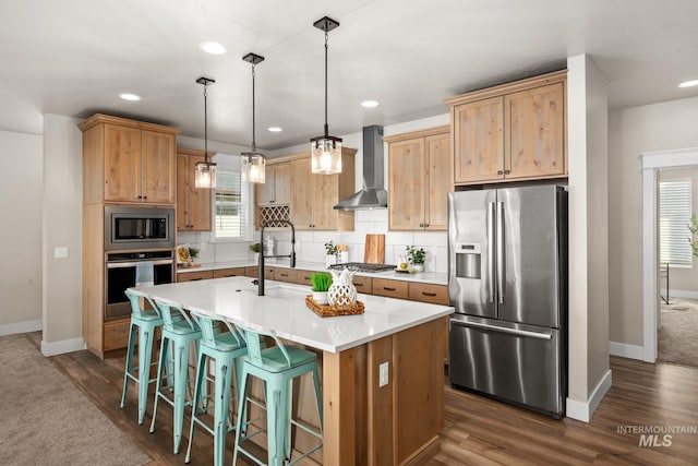 kitchen featuring wall chimney exhaust hood, a center island with sink, appliances with stainless steel finishes, decorative light fixtures, and dark hardwood / wood-style flooring