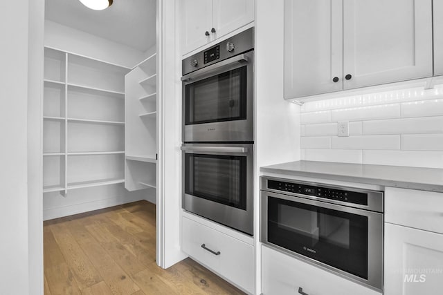 kitchen with stainless steel double oven, light wood-style floors, white cabinets, light countertops, and tasteful backsplash