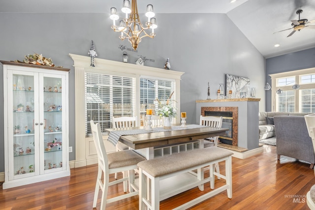 dining room with ceiling fan with notable chandelier, high vaulted ceiling, wood finished floors, and a high end fireplace