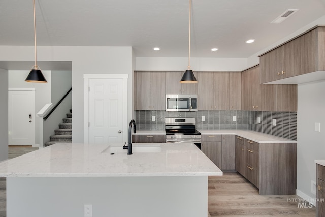 kitchen with light stone counters, an island with sink, light hardwood / wood-style floors, pendant lighting, and appliances with stainless steel finishes