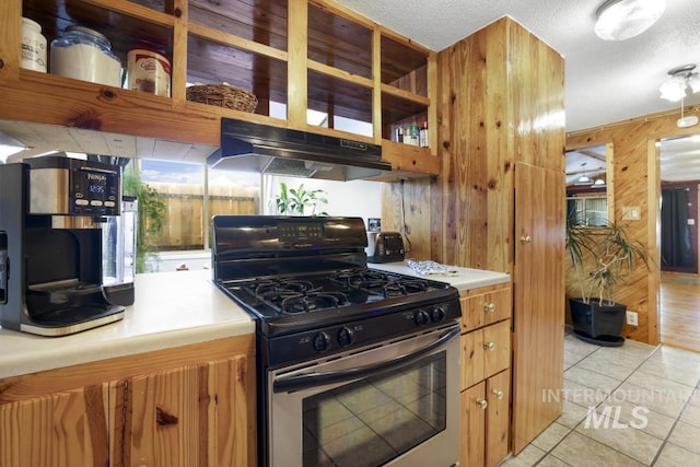 kitchen featuring stainless steel gas range oven, open shelves, wooden walls, under cabinet range hood, and light tile patterned flooring