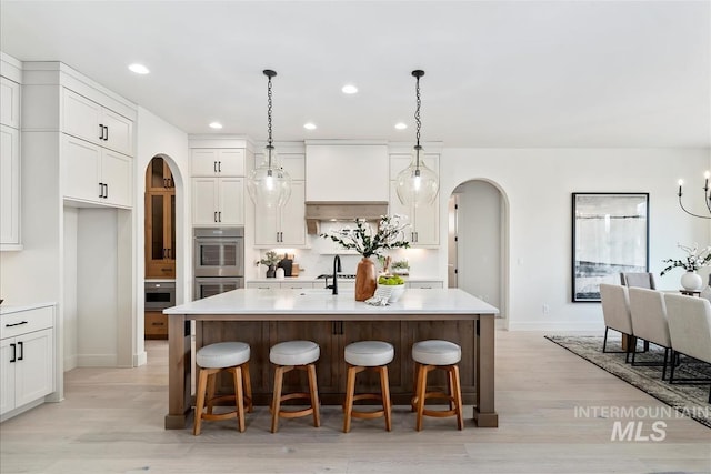 kitchen featuring arched walkways, white cabinets, an island with sink, light countertops, and double oven