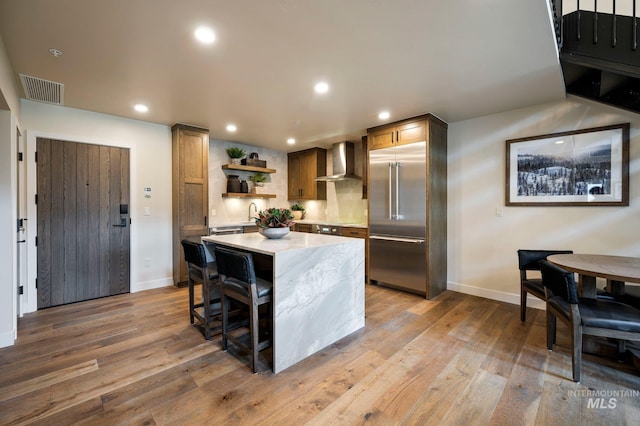 kitchen with a kitchen bar, built in refrigerator, light hardwood / wood-style flooring, a kitchen island, and wall chimney range hood