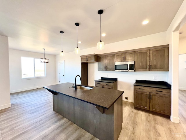 kitchen featuring dark countertops, stainless steel microwave, a kitchen island with sink, light wood-style floors, and a sink