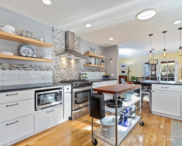 kitchen featuring stainless steel appliances, wall chimney range hood, light wood-type flooring, open shelves, and dark countertops