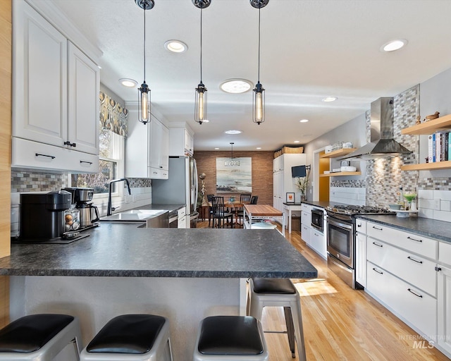 kitchen featuring dark countertops, appliances with stainless steel finishes, wall chimney range hood, open shelves, and a sink