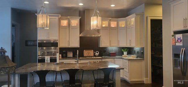 kitchen featuring an island with sink, appliances with stainless steel finishes, wall chimney range hood, and decorative light fixtures