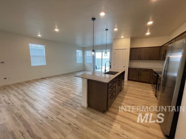 kitchen with a center island with sink, sink, hanging light fixtures, stainless steel fridge, and light stone counters