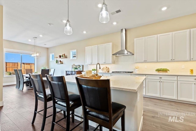 kitchen featuring visible vents, an island with sink, wall chimney range hood, stovetop, and light wood finished floors