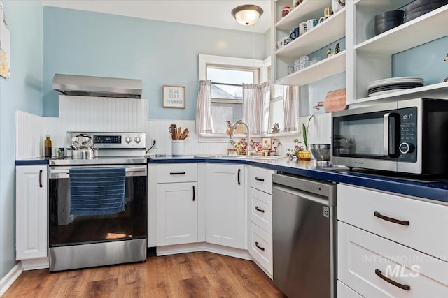 kitchen featuring a sink, wood finished floors, stainless steel appliances, white cabinets, and wall chimney range hood