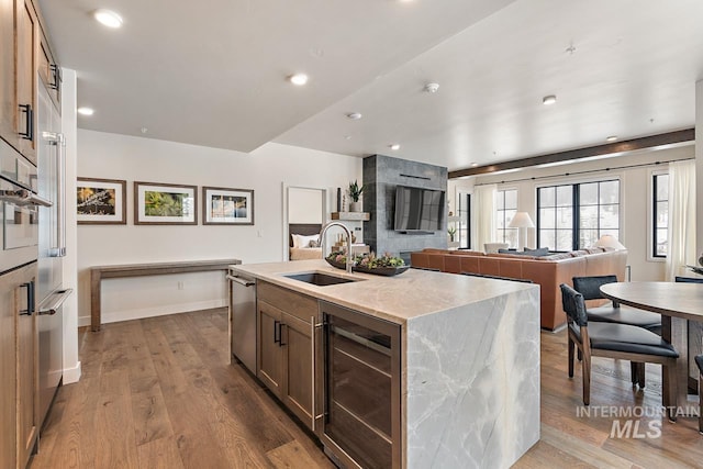 kitchen featuring sink, a kitchen island with sink, light stone counters, beverage cooler, and light wood-type flooring