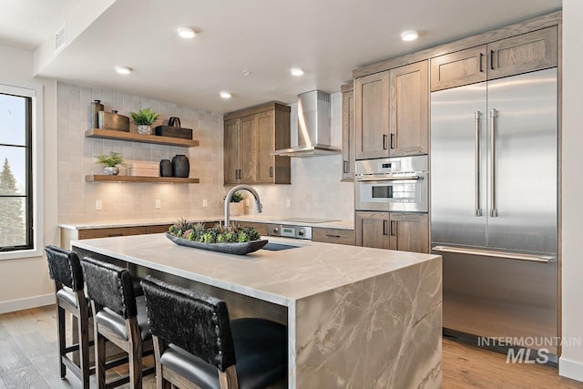 kitchen featuring appliances with stainless steel finishes, a breakfast bar, a center island with sink, light wood-type flooring, and wall chimney exhaust hood